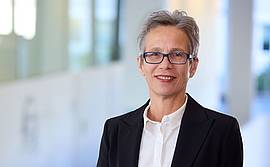Portrait einer Dame mit kurzem Haar und Brille in weißer Bluse und dunkelblauem Blazer vor Besprechungstisch
