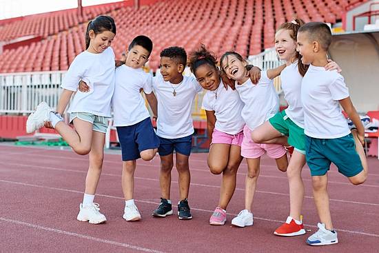 Eine Gruppe von Kindern in Sportkleidung auf einer Rennstrecke stehen in einer Reihe und haben die Arme über die Schultern der jeweils benachbarten Person gelegt, sodass sie eine Kette bilden.