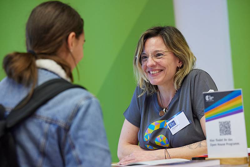 Eine Person mit T-Shirt der Hochschule Campus Wien an einem Stehtisch im Gespräch mit einer zweite Person, deren Rückseite verschwommen im Vordergrund zu sehen ist.