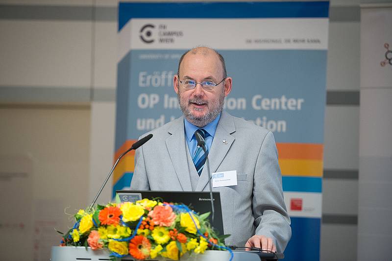 Herr mit Brille und grauem Anzug hinter Rednerpult mit Blumenbouquet im Vordergrund