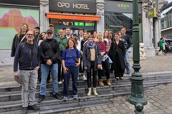 Gruppenfoto einer Exkursionsgruppe im städtischen Raum: Rund 25 Personen stehen gemeinsam auf den Stufen vor einem Gebäude und blicken in die Kamera. Im Hintergrund sind Geschäftslokale und die Fassade eines Hotels zu sehen. Die Teilnehmenden tragen Freizeitkleidung und wirken entspannt. Die Aufnahme entstand bei bewölktem Wetter in einer europäischen Innenstadt.