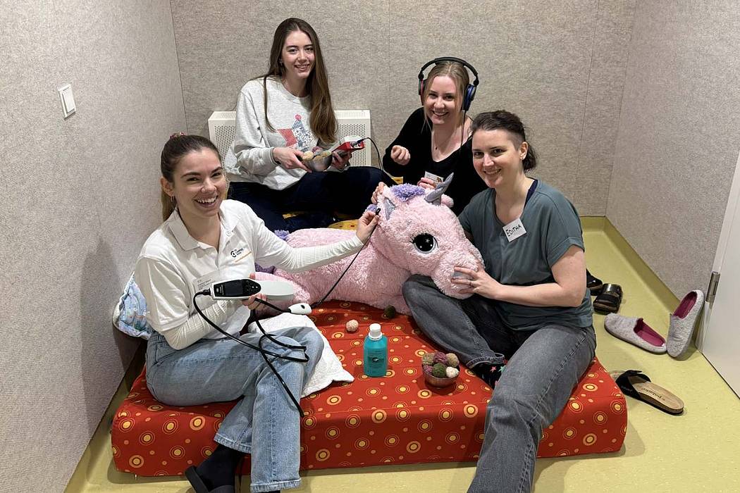 Vier Personen sitzen in einem kleinen, ruhigen Raum auf Bodenkissen und beschäftigen sich gemeinsam mit einem großen rosafarbenen Stofftier sowie Kopfhörern und technischen Geräten.