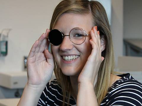 Studierende trägt ein Instrument in der Hand, in der Form von einer Brillenfassung mit Bügel, die linke Seite ist schwarz abgedeckt. Sie lächelt in die Kamera und hält mit den Händen die Bügel fest.