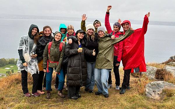 © privat Eine Gruppe von zehn Personen steht auf einer grasbewachsenen Anhöhe mit Felsen, im Hintergrund ist ein großer See unter grauem, wolkenverhangenem Himmel zu sehen. Die Menschen tragen wetterfeste Kleidung wie Regenjacken, Ponchos und Mützen. Einige haben Rucksäcke, andere halten Getränke. Mehrere Personen heben die Arme fröhlich in die Luft, die Stimmung wirkt ausgelassen und gemeinschaftlich