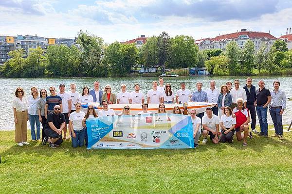 © HCW/Schedl Gruppenfoto des Betonkanu-Teams der Hochschule Campus Wien an einem Flussufer. Rund 30 Personen stehen und knien gemeinsam auf einer Wiese hinter einem Banner mit Logos und der Aufschrift „HCW Betonkanu“. Im Hintergrund sind Wasser, Bäume und Gebäude zu sehen.