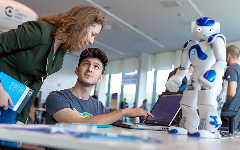 Zwei Personen an einem tisch auf dem ein geöffneter Laptop neben einem etwa 20cm großen humanoiden Roboter steht