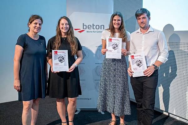 © Beton Dialog Vier Personen stehen nebeneinander auf einer Bühne vor einem Roll-up von Beton Dialog Österreich. Drei von ihnen halten Urkunden der Concrete Design Competition in den Händen und blicken lächelnd in die Kamera. Die Gruppe trägt sommerliche, schlichte Kleidung und posiert für ein Erinnerungsfoto der Preisverleihung.