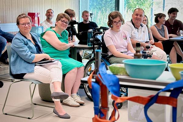 © HCW Mehrere Personen sitzen in einem hellen Seminarraum und verfolgen aufmerksam eine Präsentation. Im Vordergrund steht ein Tisch mit Schüsseln und bunten Hebegurten, im Publikum sitzt unter anderem eine Person im Rollstuhl. Die Teilnehmenden blicken nach vorne und wirken interessiert.