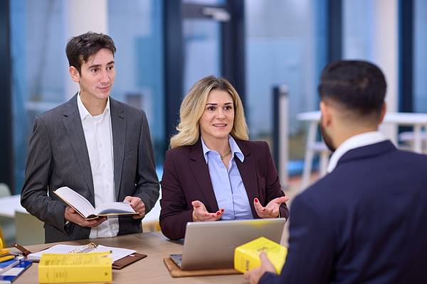 Drei Personen stehen an einem Tisch. Auf dem Tisch liegen Bücher und ein Laptop.