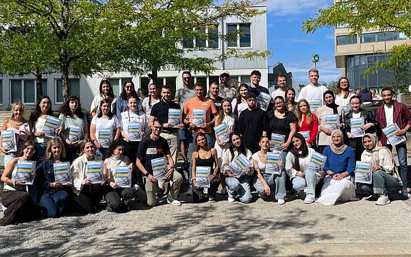Eine große Gruppe junger Menschen posiert auf Stiegen, sie halten bunte Mappen in der Hand. Im Hintergrund ist ein Gebäude sowie Himmel zu sehen.