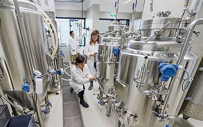 © HCW | Schedl Drei Personen in Laborkitteln arbeiten inmitten von Edelstahl-Gärtanks