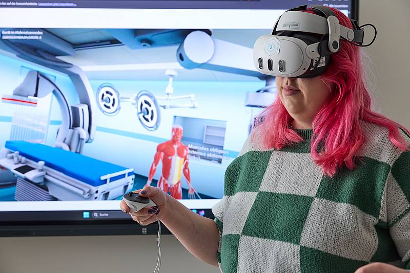 Eine Person mit VR-Brille und einer Kontrolleinheit in der Hand. Im Hintergrund ein Monitor, der eine Simulation in einem Patientenzimmer zeigt.