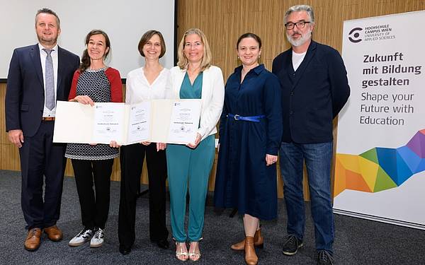 Gruppenfoto bei einer feierlichen Urkundenverleihung an der Hochschule Campus Wien. Drei Personen in der Mitte halten Zertifikate, flankiert von zwei weiteren Personen. Rechts im Bild steht ein Roll-up mit dem Logo der Hochschule