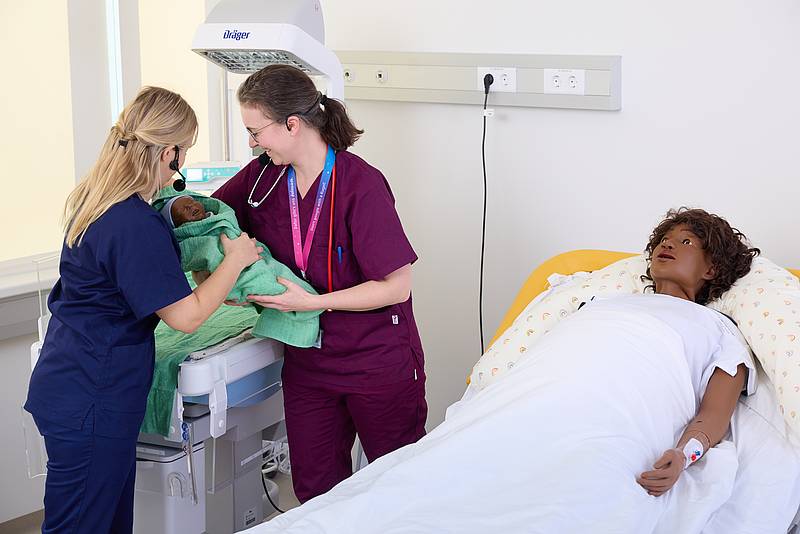 Zwei Personen in Kasaks in einem Krankenzimmer halten ein Modell eines Säuglings. Daneben ein Krankenbett mit einer Puppe, die eine Patientin darstellt.
