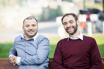 Herr in hellblauem Hemd sitzt neben anderem Herrn in dunkelrotem Pullover auf einer Bank im Hintergrund eine Wiese und eine Straße