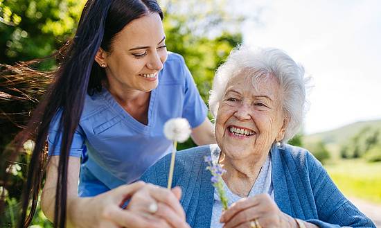 Eine junge Person reicht einer älteren, sitzenden Person eine Pusteblume, beide lächeln