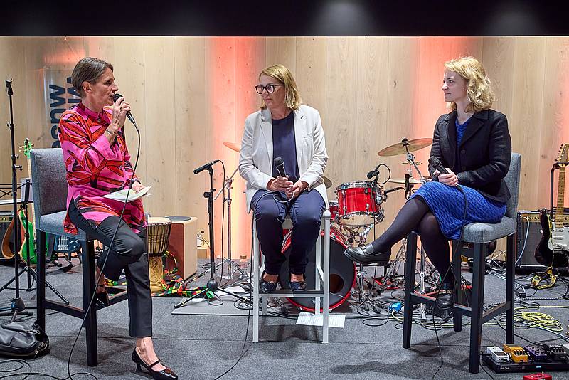 Drei Personen bei einer Diskussion auf einer Bühne. Im Hintergrund Band-Equipment