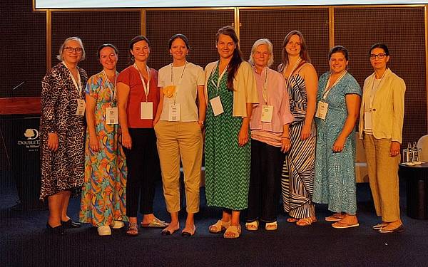 Neun Personen stehen gemeinsam auf einer Bühne bei einer Konferenz, alle tragen Namensschilder und sommerliche Kleidung