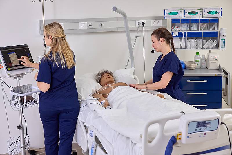Zwei Studierende an einem Krankenbett mit einer Patientenpuppe und einem Bildschirm, der die simulierten Vitalwerte zeigt.