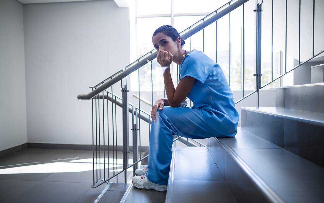 © iStock.com / Wavebreakmedia Eine Pflegekraft sitzt allein in einem Stiegenhaus, das Kinn in die Hand gestützt