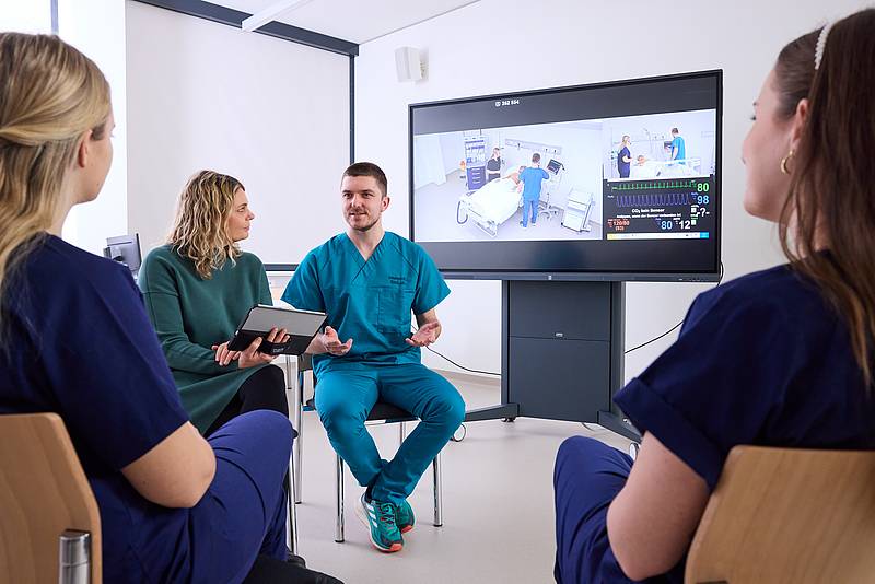 Vier Personen in einem kleinen Stuhlkreis, die miteinander sprechen. Im Hintergrund ein großer Monitor, der Aufnahmen aus verschiedenen Patientenzimmern zeigt.