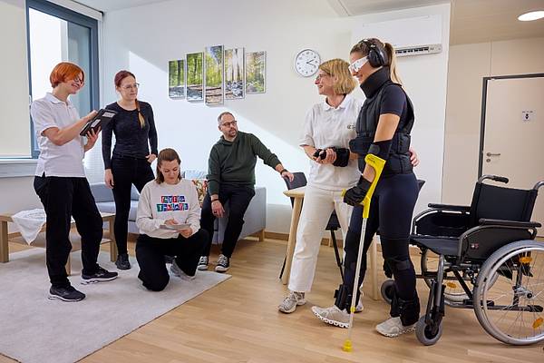 © HCW/Schedl In einem hellen Therapieraum üben mehrere Personen gemeinsam das Gehen. Eine Patientin mit Augenbinde und Trainingsausrüstung wird dabei von einer Therapeutin begleitet. Zwei weitere Therapeutinnen sowie drei Personen beobachten und unterstützen die Übung. Neben der Patientin stehen ein Gehstock und ein Rollstuhl.