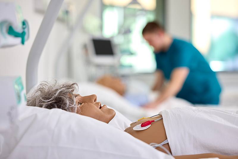 Im Vordergrund eine Patientenpuppe in einem Krankenbett. Im Hintergrund eine Person im Kasak über ein zweites Krankenbett gebeugt.