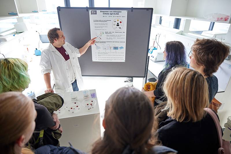 Eine Person steht in einem Labor vor einem Plakat zu Allergien. Vor ihm befinden sich Chemie-Behältnisse und eine Gruppe Interessierter.