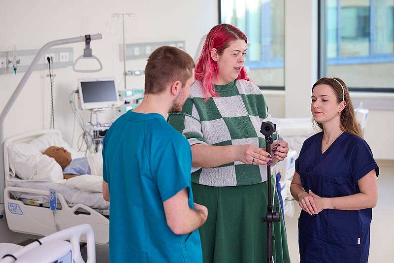 Drei Personen in einem Patientenzimmer sprechen miteinander. Im Hintergrund eine Patientenpuppe in einem Bett.
