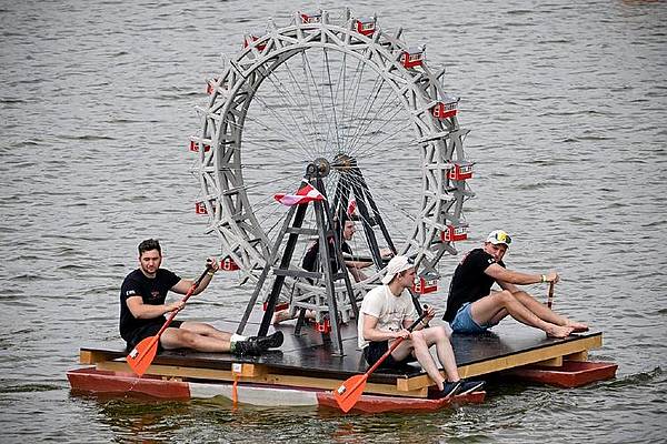 Vier Personen sitzen auf einem schwimmenden Floß und paddeln auf einem Gewässer. In der Mitte des Floßes befindet sich ein großes, detailreich nachgebildetes Wiener Riesenrad aus Beton. Die Konstruktion dient als ungewöhnliches Wasserfahrzeug und ist Teil eines Wettbewerbs. Im Hintergrund ist die Wasseroberfläche zu sehen.