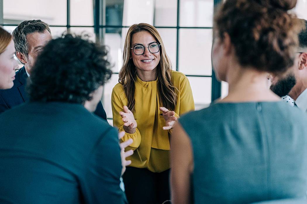 © VioletaStoimenova/iStock.com Frau mit Bille und gelben Oberteil erklärt anderen Personen etwas