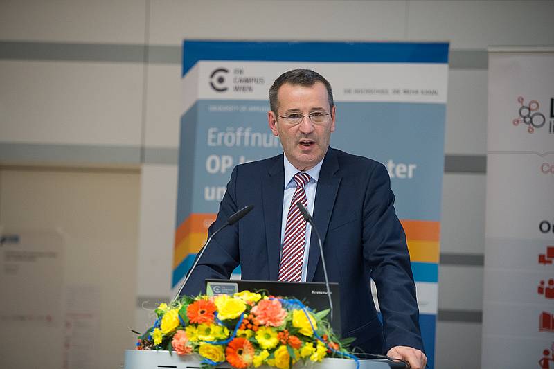 Herr mit Brille in dunkelblauem Anzug hinter Rednerpult mit Blumenbouquet im Vordergrund