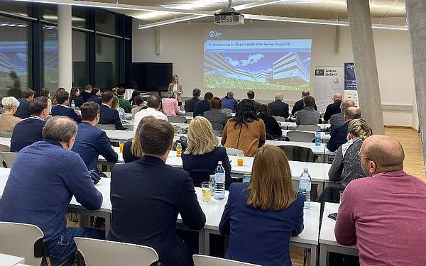 Ein Lehrsaal von hinten, vorne steht eine Person vor einer Leinwand und haltet einen Vortrag