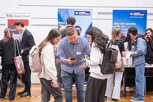 Zwei Besucher*innen der Karrieremesse Technik tauschen mit Mitarbeiter eines Unternehmens Kontaktdaten aus