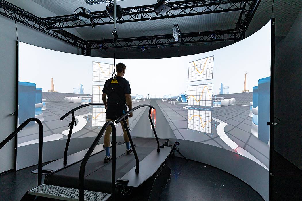 Mann von hinten geht auf einem Laufband, vor ihm ein virtueller Hintergrund auf einer 180-Grad-Leinwand