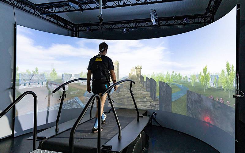Ansicht von hinten. Eine Person im Ganglabor der HCW auf einem Laufband. Vor der Person befindet sich eine große Bildschirmwand, die eine Landschaft zeigt