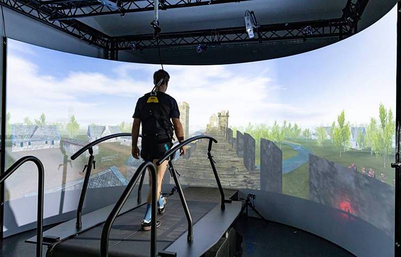 Ansicht von hinten. Eine Person im Ganglabor der HCW auf einem Laufband. Vor der Person befindet sich eine große Bildschirmwand, die eine Landschaft zeigt