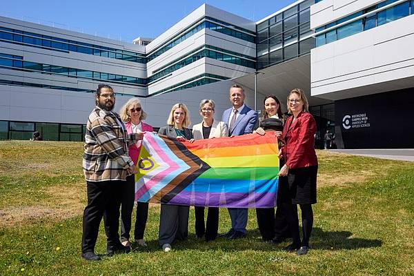 Sechs Personen stehen vor dem Gebäude der Hochschule Campus Wien auf einer Wiese und halten gemeinsam eine Progress-Pride-Flagge. Im Hintergrund ist die moderne Hochschulfassade bei sonnigem Wetter zu sehen. Die Gruppe blickt in die Kamera und setzt ein sichtbares Zeichen für Vielfalt und Inklusion.