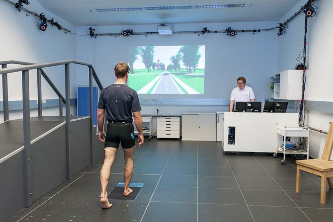 Studierender betritt einen schwarzen Plattenbereich im Boden. Vor ihm befindet sich eine Leinwand, auf der eine Straße angezeigt wird. Neben der Leinwand ist ein Tisch, auf dem sich ein Computer und mehreren Monitoren befinden. Dort steht eine Person und beobachtet die Bewegungen des Studierenden