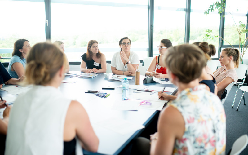 ©Ulrich Dobiasch Eine Gruppe von Frauen sitzt zusammen an einem Tisch und diskutieren. Der Raum ist hell und große Fenster sind hinter den Damen zu sehen. Auf dem Tisch stehen Wasserflaschen und Broschüren und Stifte liegen verstreut auf der Tischplatte.