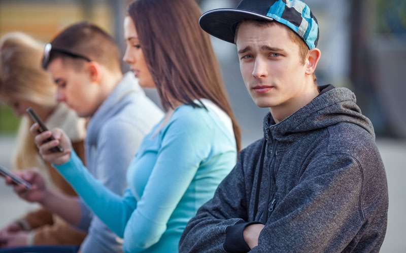 Jugendlicher mit Kappe im Vordergrund, der in die Kamera sieht, 3 Jugendliche im Hintergrund, die ins Handy schauen