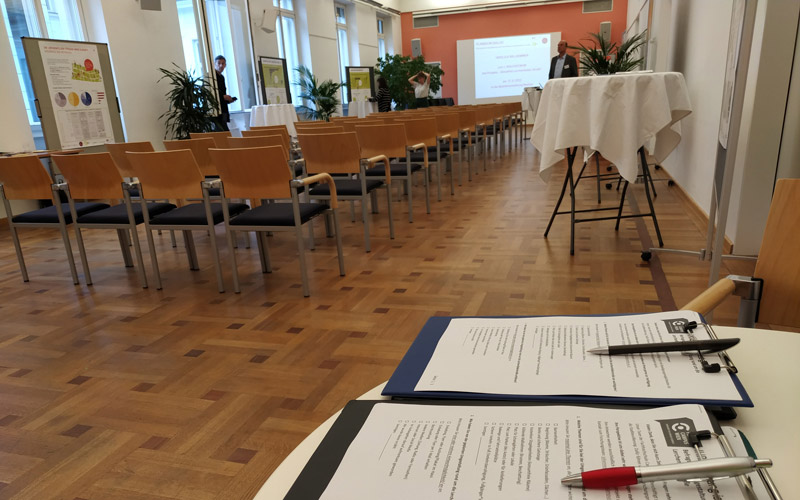 © HCW Ein leerer Präsentationssaal ist abgebildet, im Vordergrund liegen zwei Formulare zum ausfüllen