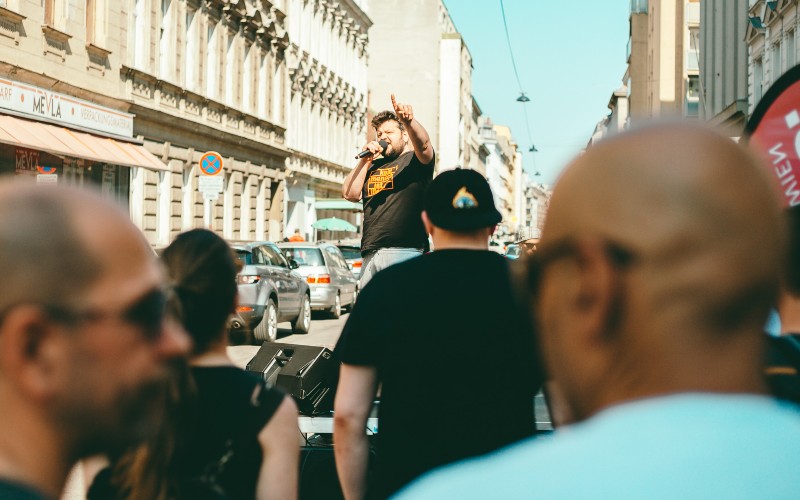 Ein Mann spricht durch ein Mikrofon zu einer Menge und streckt den Zeigefinger gen Himmel