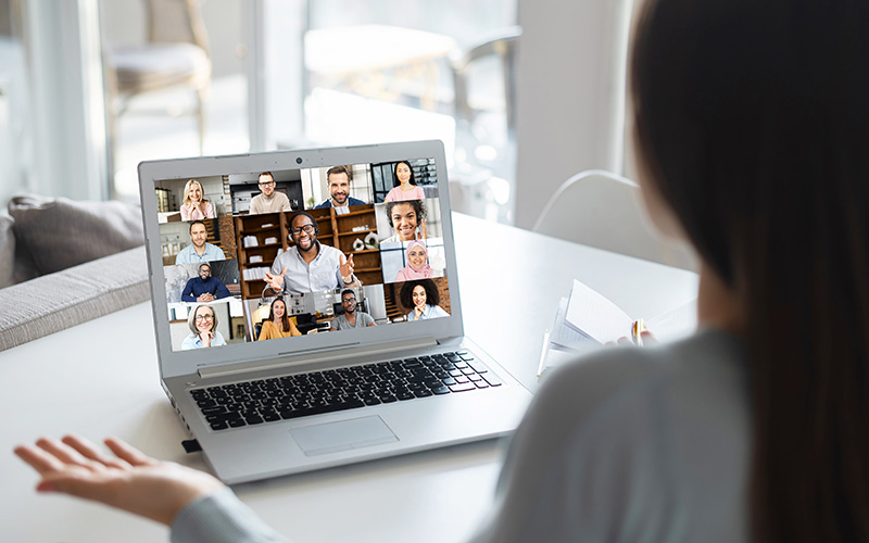 Eine Person sitzt und gestikuliert vor einem Laptop, auf dem eine Videokonferenz mit 13 Personen angezeigt wird