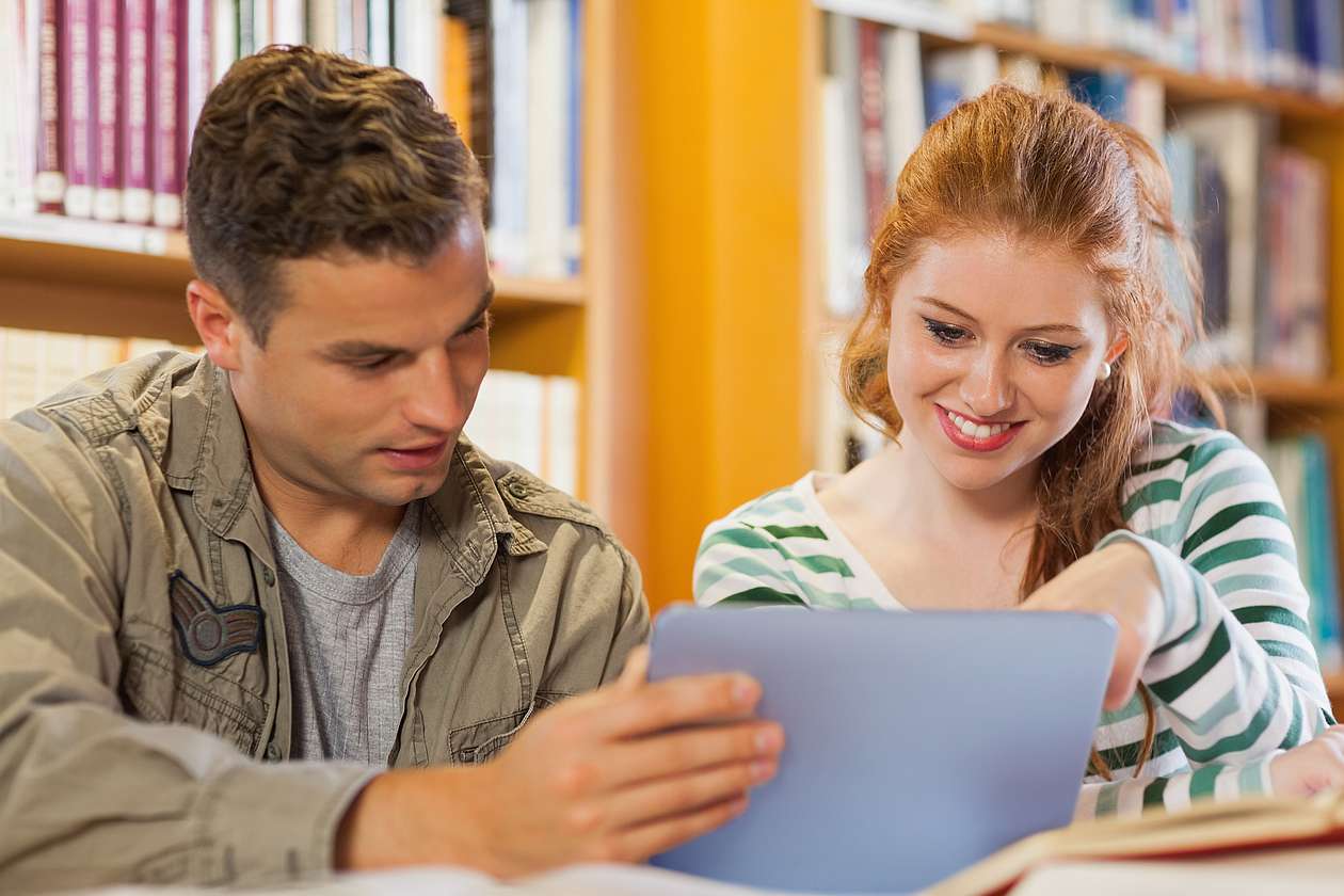 Zwei Studierende in einer Bibliothek schauen lächelnd gemeinsam auf ein Tablet
