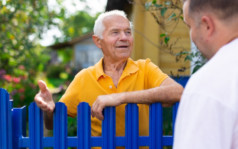© Caftor/Shutterstock Ein Senior lehnt sich auf einen Gartenzaun um mit seinem Nachbar zu sprechen