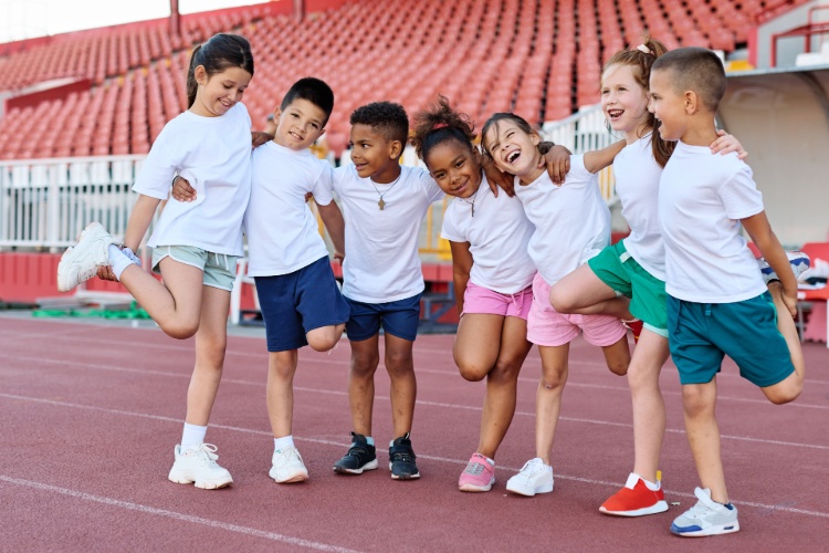 Eine Gruppe von Kindern in Sportkleidung auf einer Rennstrecke stehen in einer Reihe und haben die Arme über die Schultern der jeweils benachbarten Person gelegt, sodass sie eine Kette bilden.