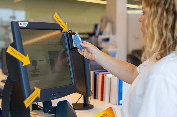 Ein Computerbildschirm mit aufgeklebten Pfeilen, die zeigen, wo man den Bibliotheksausweis zum Scannen anhalten muss, um selbständig Bücher zu entlehnen.