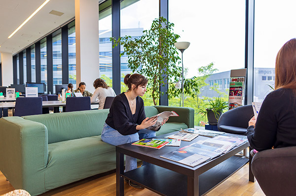 Bild einer Sitzecke mit Couch und niedrigem Glastisch. Dahinter sieht man mehrere Tischreihen mit PC-Arbeitsplätzen.