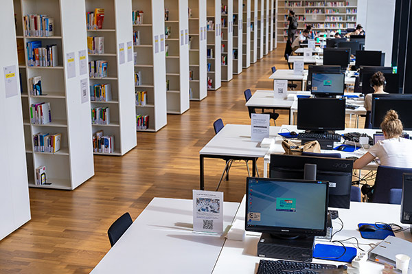 Links eine lange Reihe von Bücherregalen; rechts mehrere Reihen mit PC-Arbeitsplätzen